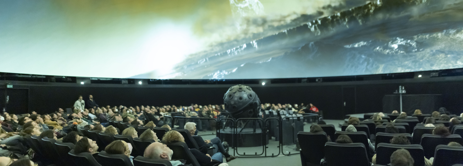 A picture of people in a planetarium, watching a screen in front and above them.