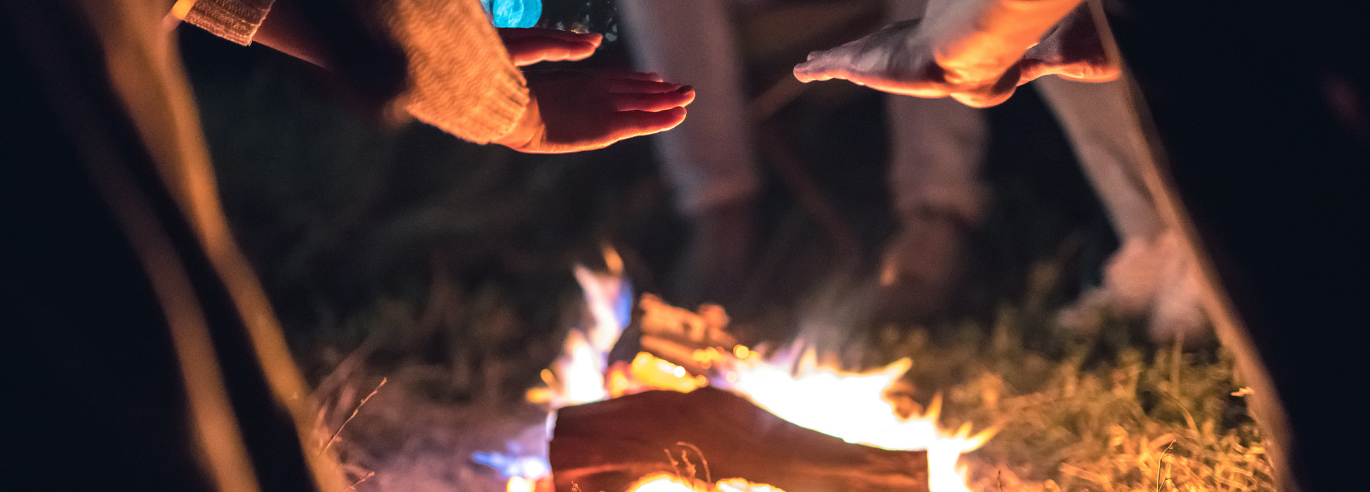 This image captures a cozy outdoor scene where people are gathered around a small campfire at night. Several hands are extended toward the fire, likely to warm up, as the flames crackle over the logs. The soft glow of the fire illuminates the surrounding area, casting warm light on the hands and nearby grass.