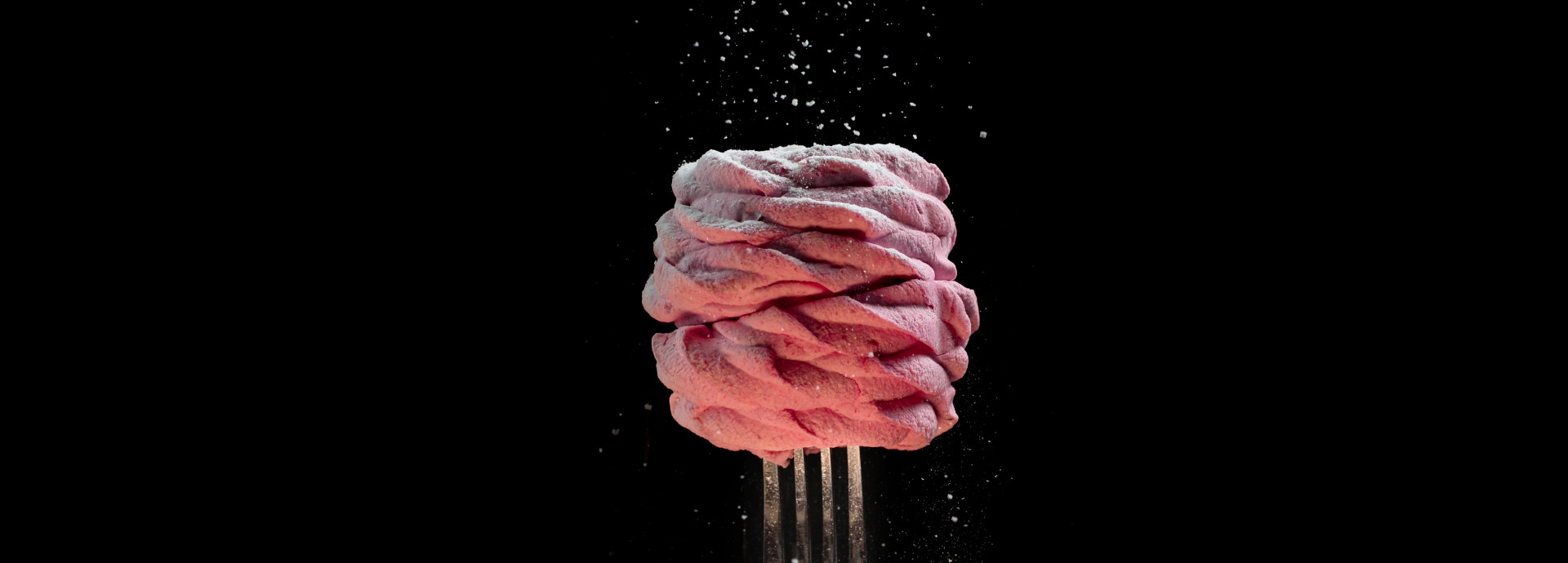 The image depicts a visually striking food item, likely a dessert, against a black background. The dessert consists of twisted pink meringue or a similar pastry, reminding human brain structure, sitting atop a fork that is held upright. Powdered sugar is being sprinkled over the dessert, creating a delicate dusting effect, with fine particles of sugar visible in mid-air. The use of contrasting dark and light elements, as well as the minimalistic setup, gives the image a refined, artistic feel.