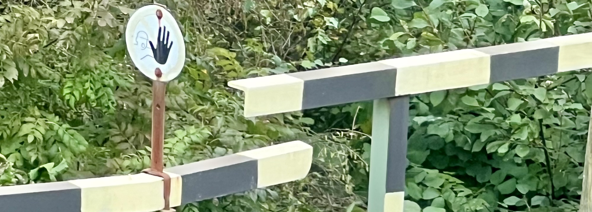 The image shows a barrier gate, likely at a road or railway crossing, painted in alternating black and white stripes. Next to the gate is a circular sign with a handprint design, suggesting a stop or caution signal. The background is filled with dense green foliage, indicating a natural or rural setting. The design of the gate and the sign imply a request to halt, possibly to indicate restricted access or a need for caution at the crossing.