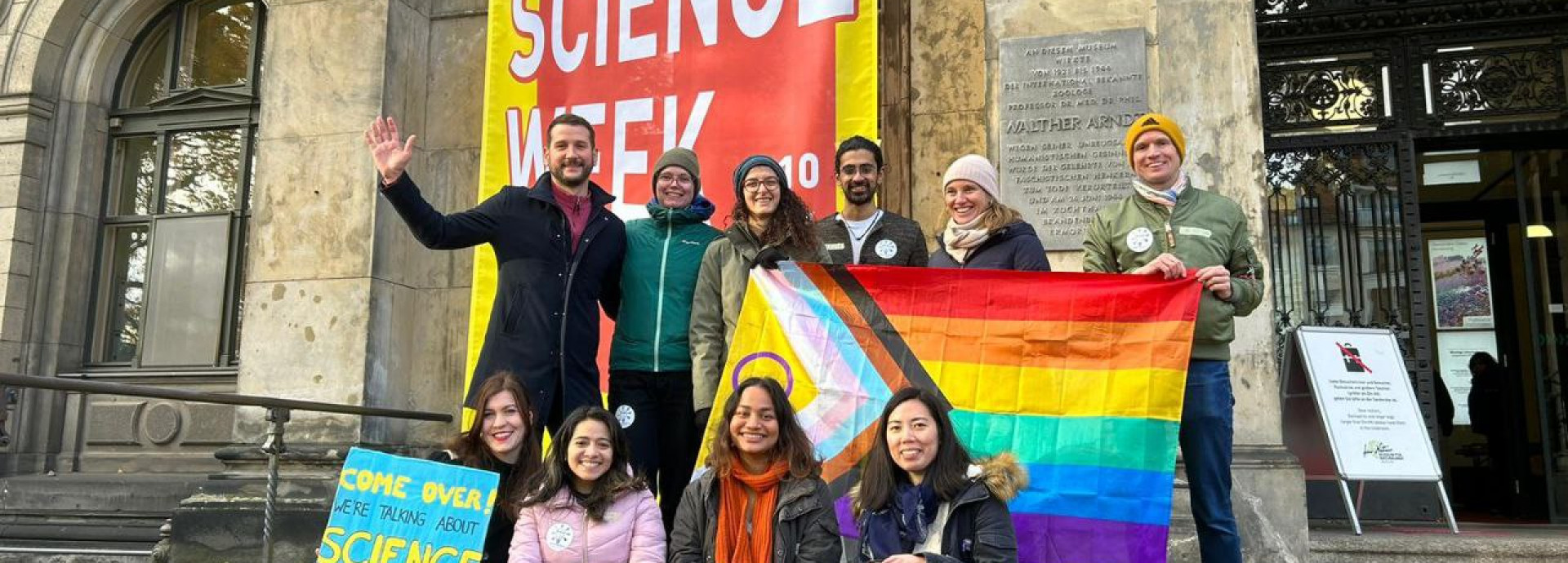 Soapbox Science Berlin and LGBTQ+ STEM Berlin teams pose with pride flag in front of Museum für Naturkunde.