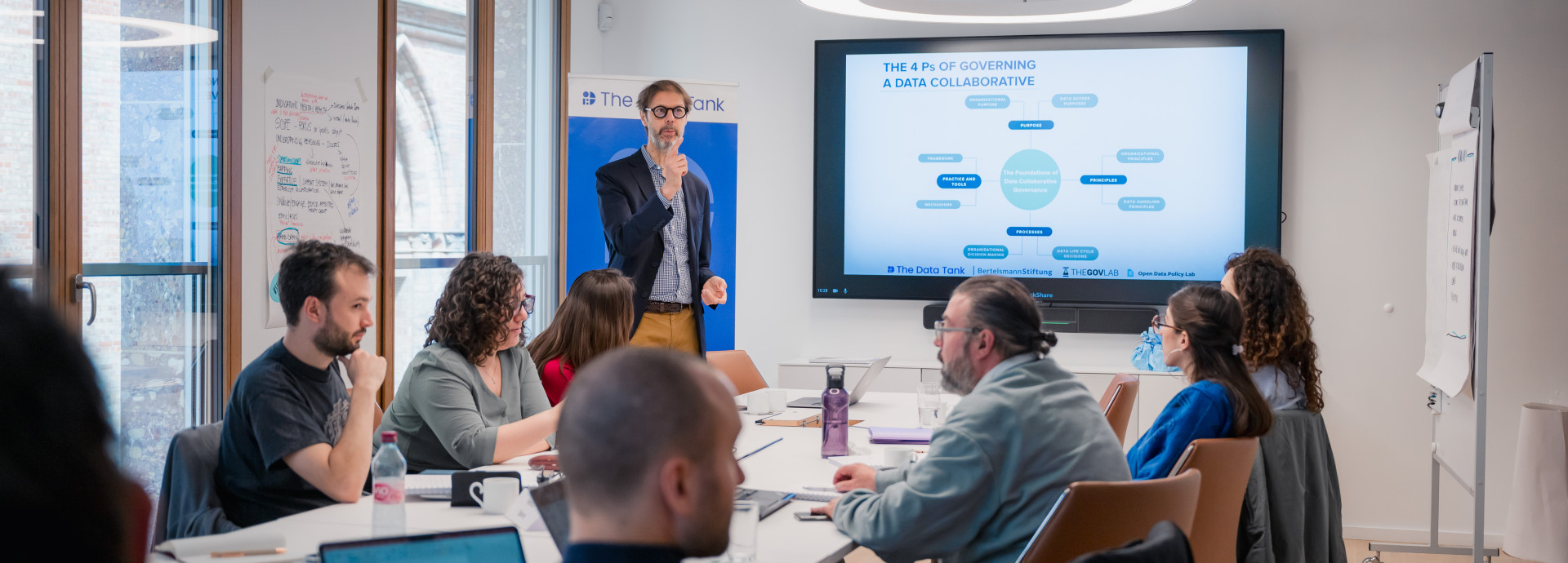 This image shows a meeting or workshop taking place in a modern conference room. Around a large table, several individuals are seated with laptops, notebooks, and coffee cups, actively listening to a speaker standing in front of a presentation screen. The speaker is holding a microphone and gesturing, explaining a slide titled "The 4 Ps of Governing a Data Collaborative." The slide contains a flowchart or diagram with various text bubbles, likely outlining the framework being discussed. 