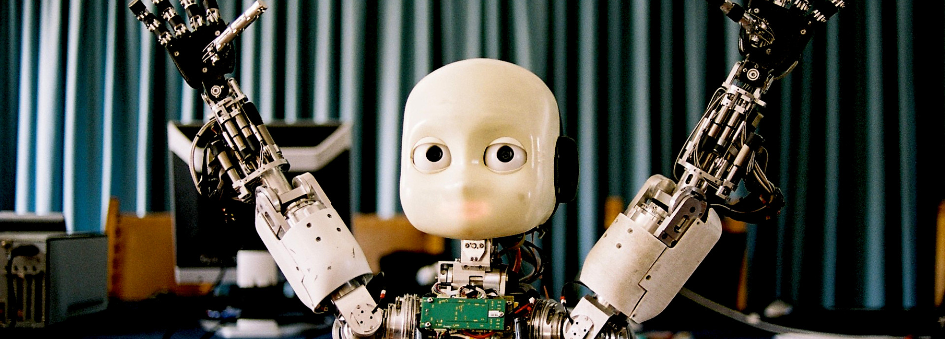 Das Bild zeigt einen humanoiden Roboter mit einem minimalistischen Gesicht, großen Augen und erhobenen Armen. Der Roboter hat komplexe mechanische Hände mit vielen beweglichen Teilen und ist teilweise freigelegt, sodass seine inneren elektronischen Komponenten sichtbar sind. Im Hintergrund hängt ein Vorhang, und es befinden sich Computer und technische Ausrüstung auf einem Tisch.