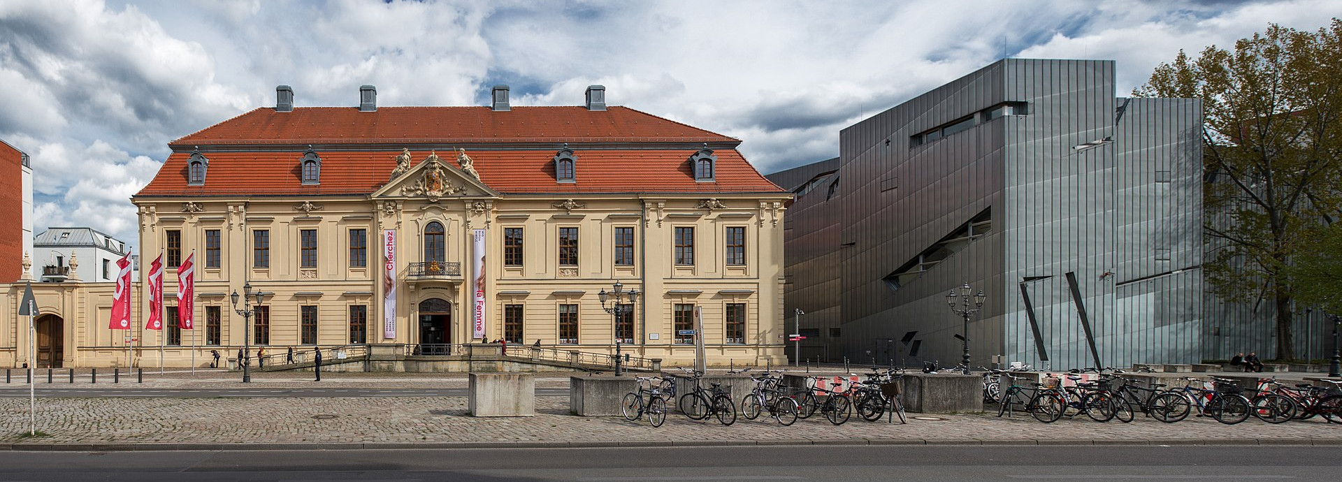 Two buildings: an old building on the left, a modern building on the right. In front there is a street.