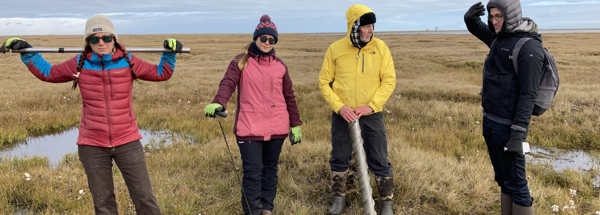 Two women and two men in bright-coloured outdoor clothing standing in the tundra with permafrost coring equipment.