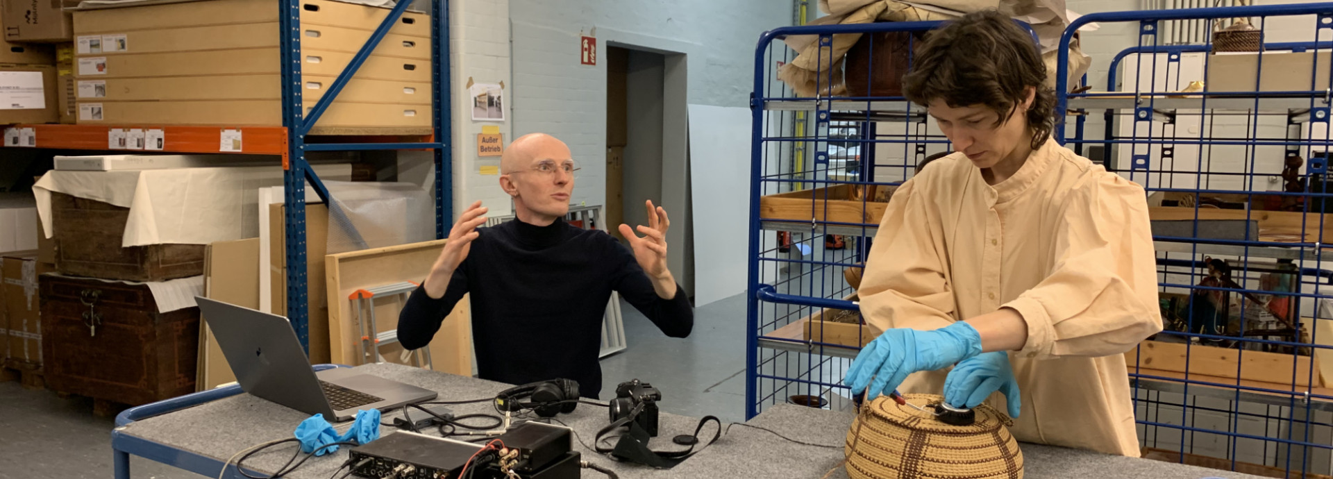 Two people are in a museum storage. One on the left side is seated and talking. One on the righ side standing and using a sound device over a museum collection item.