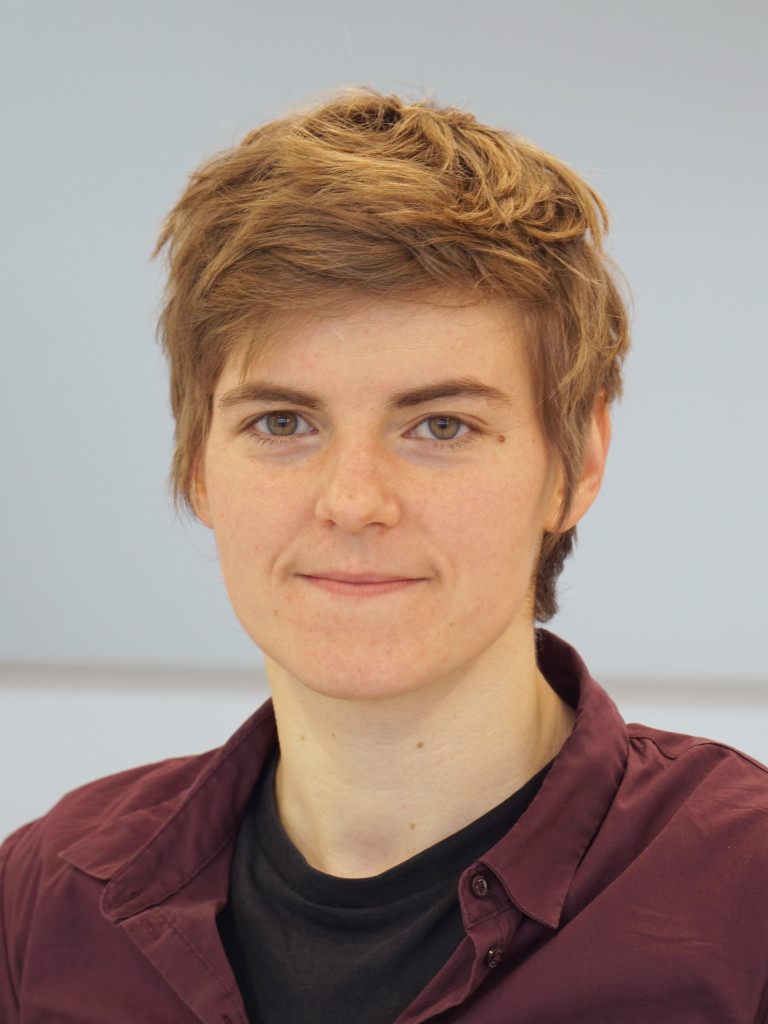 Portaitfoto von Annika. Annika trägt ein weinrotes Hemd, darunter ein schwarzes T-shirt. Das Gesicht ist im Zentrum des Bildes und Annika blickt mit einem freundlichen Gesichtsausdruck in die Kamera. Annika hat helle Haut und kurze dunkelblonde Haare.