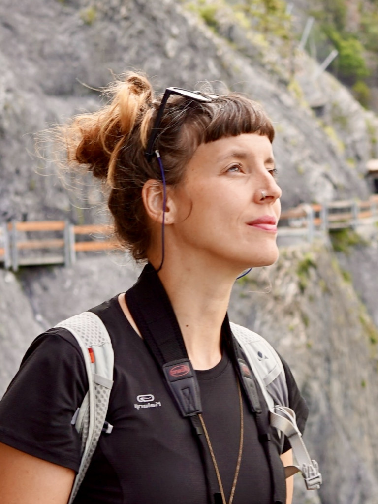 Profile of a woman with brown hair standing infront of a mountain slope looking into up into the right corner of the image.
