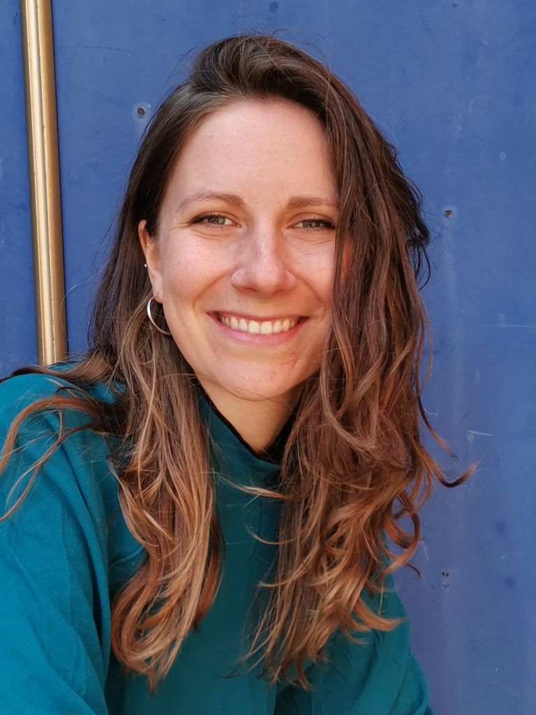Smiling young woman with long brown hair in front of a blue wall.