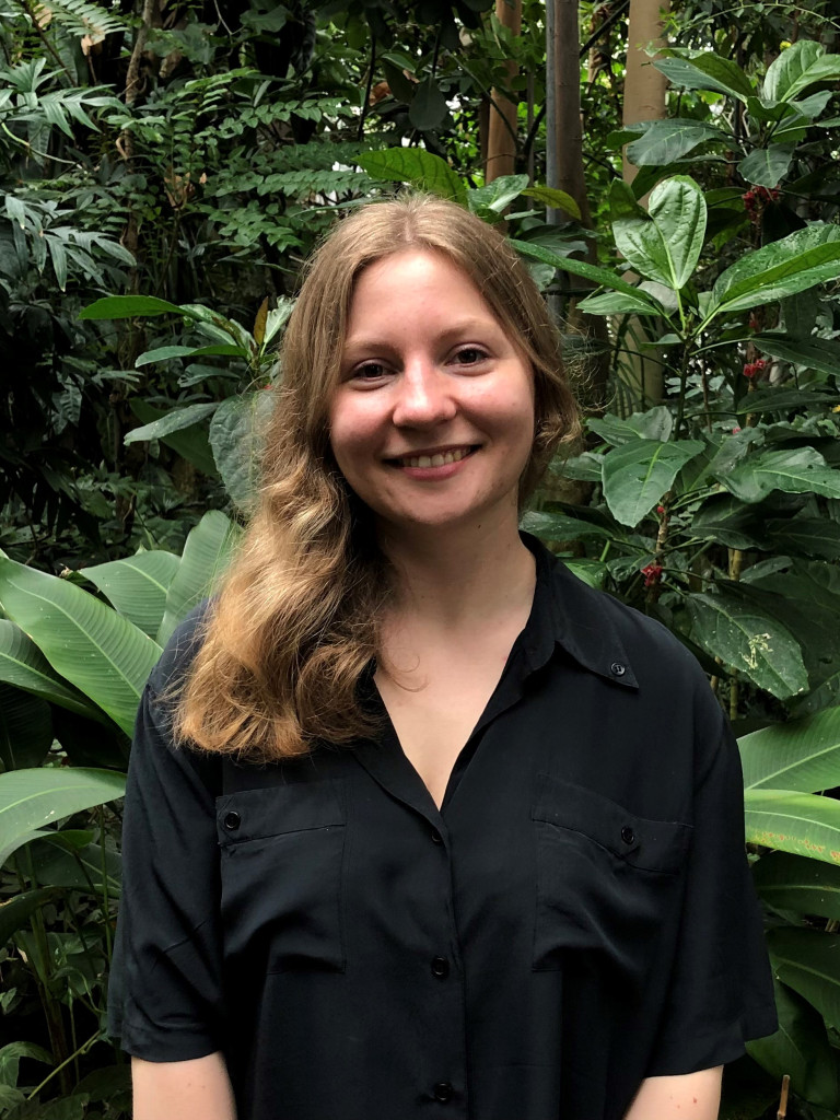 Camilla Hörsch smiles at the camera. She is wearing a black shirt and her hair on her right shoulder. In the background there are tropical plants.