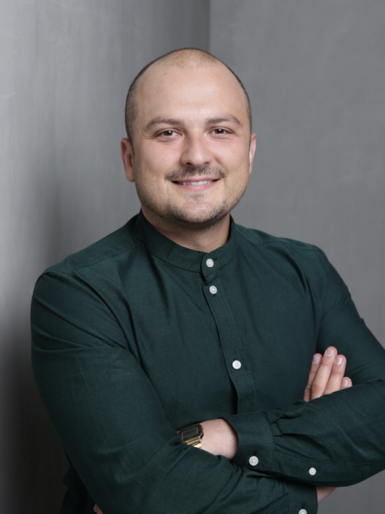 A professional shot of Mateusz Ambrozkiewicz smiling, wearing a button up shirt and with arms crossed.