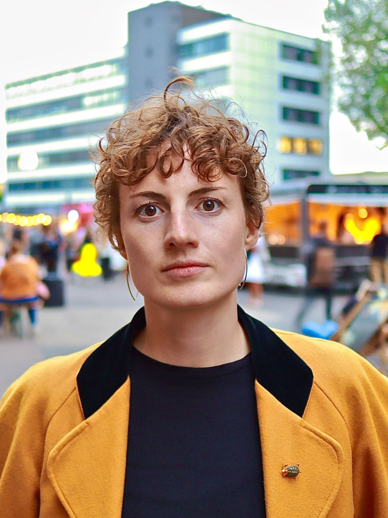 Miriam Neuhausen looks into the camera while wearing a mustard/black coat. In the background, people are eating and drinking at tables in an open space. There are buildings and trees around the plaza.