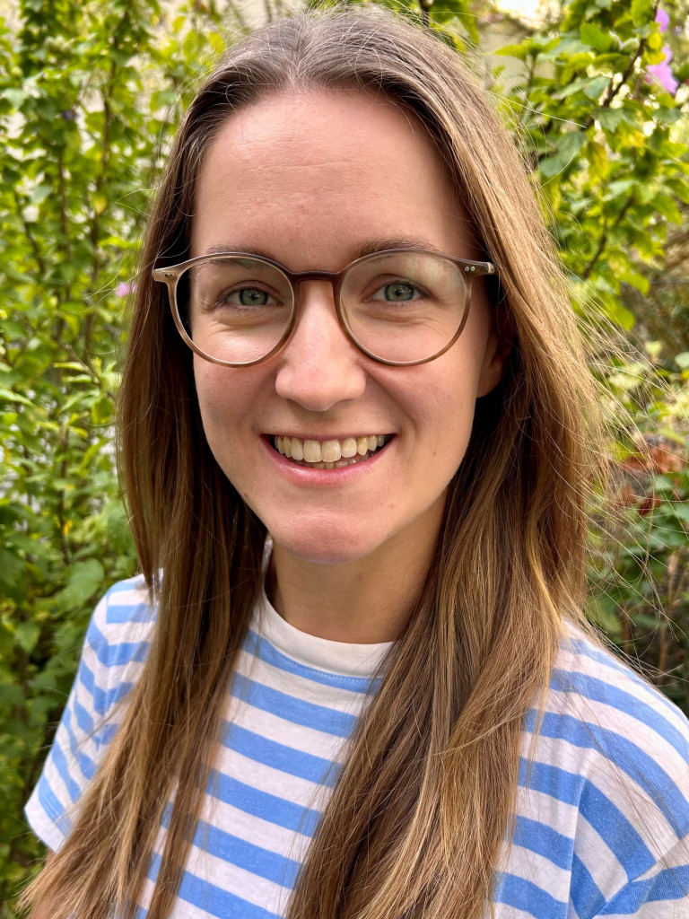Vanda Púčiková smiles at the camera. She is wearing a t-shirt with blue and white horizontal stripes, and round framed glasses. In the background, a bush with some pink flowers.