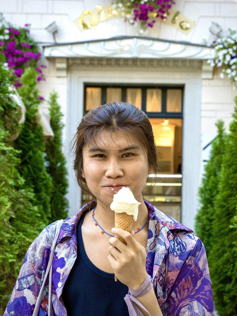 Zimai Li looks at the camera slightly smiling as he eats an ice cream. He is wearing a colourful purple shirt over a black top. In the background there is a door to an ornamented white building, and small pine trees along the sides.