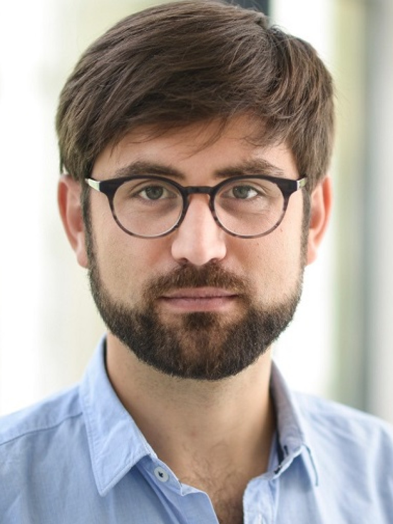 Das Bild zeigt einen Mann mit kurzen braunen Haaren, Bart und einer Brille mit dunklem Rahmen. Er trägt ein hellblaues Hemd und blickt direkt in die Kamera. Der Hintergrund ist unscharf und zeigt helle, lichtdurchflutete Fenster, was auf eine Innenaufnahme hinweist. Sein Ausdruck wirkt ruhig und konzentriert.