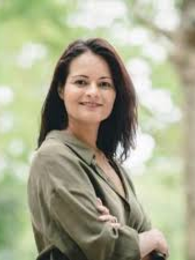 Standing woman who is smiling  with crossed arms. Background green trees.
