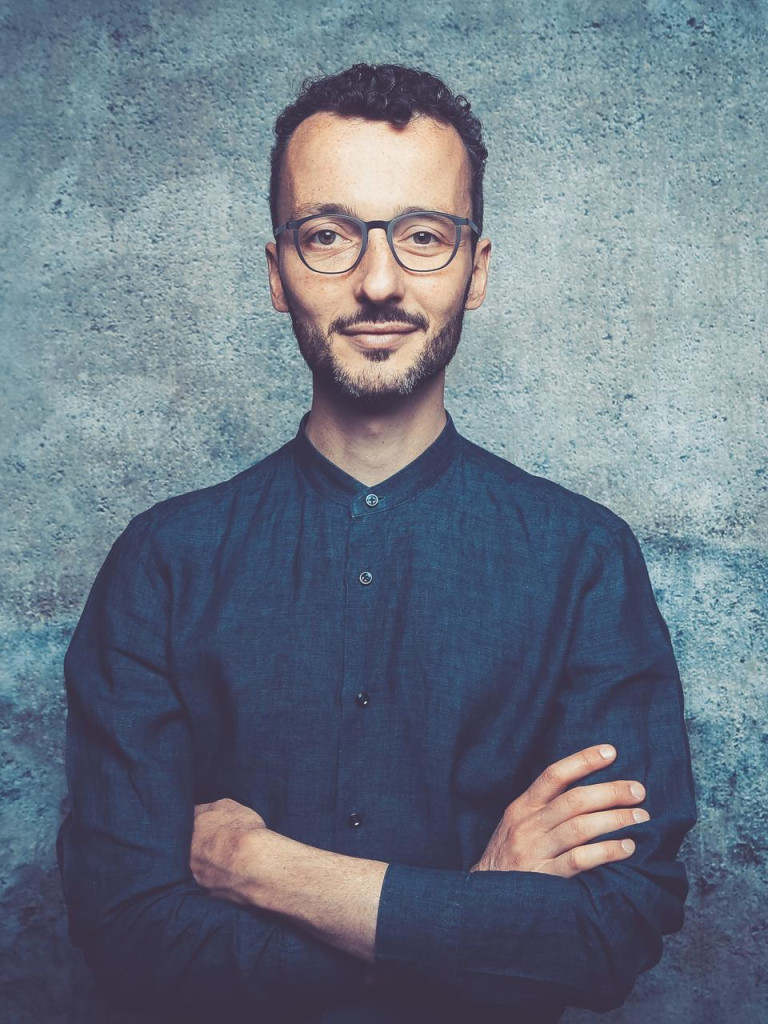 Mann mit kurzen Haaren und Brille vor blauem Hintergrund. 
