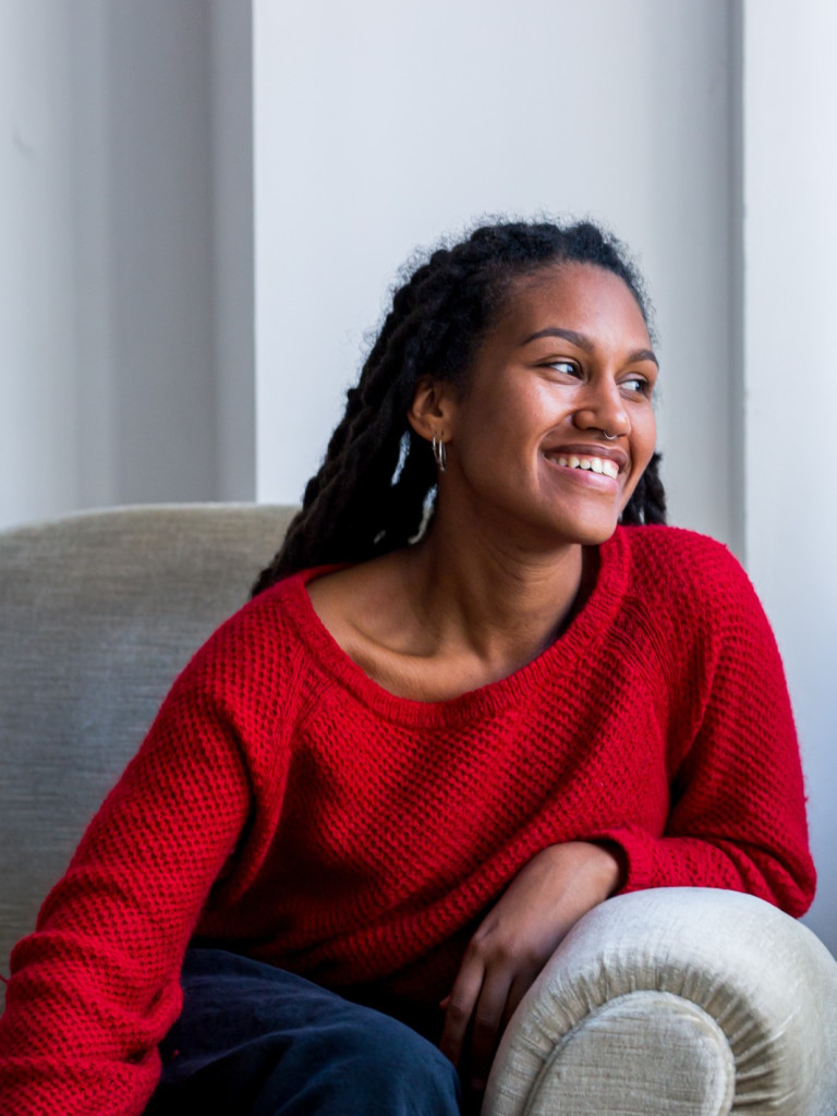 Emma-Lee Amponsah is wearing a red cardigan over blue jeans sitting on a beige couch, looking left with a smile on her face