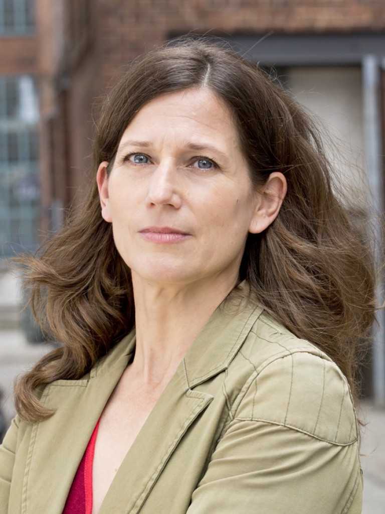 Regine, with longer dark blind hair, is standing on a courtyard and looks into the camera