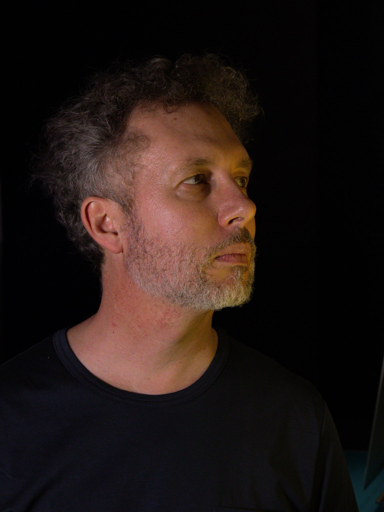 Chris is standing before black background and looks to the roght of the camera