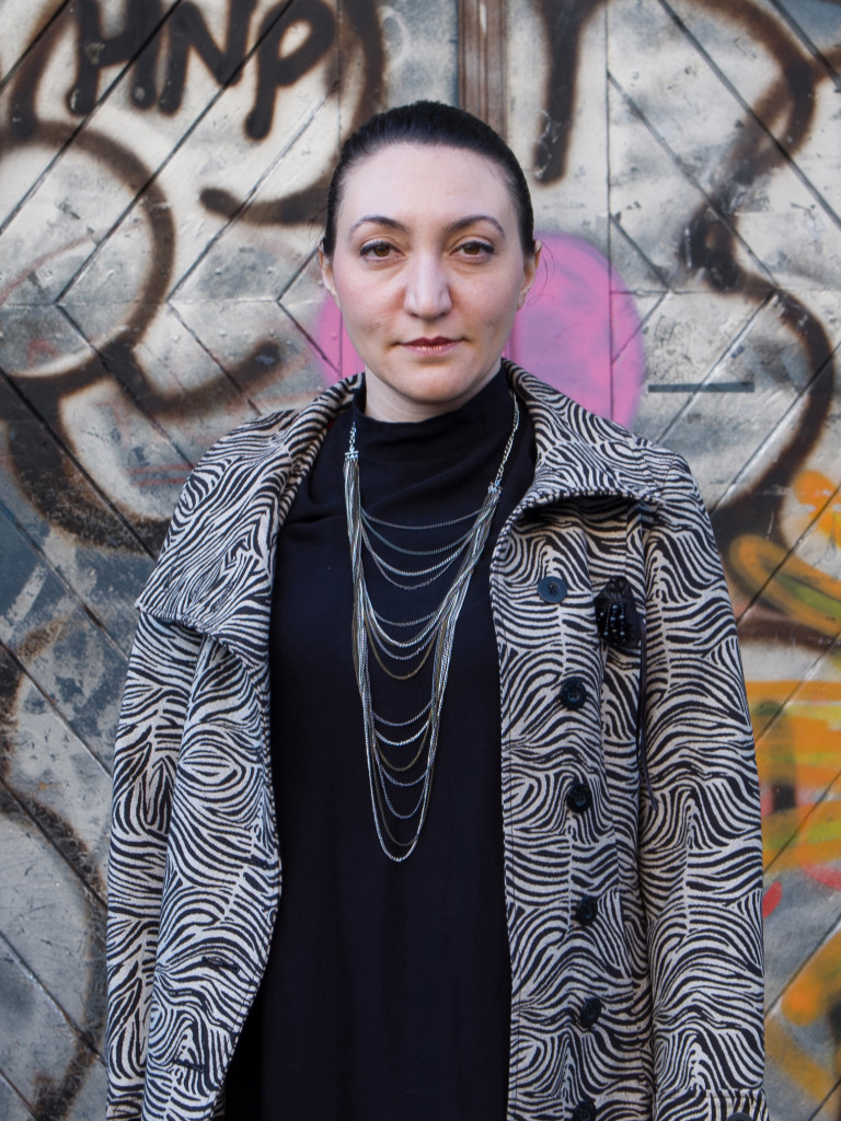 A female with black and white jacket is standing infront of a graffiti covered surface.