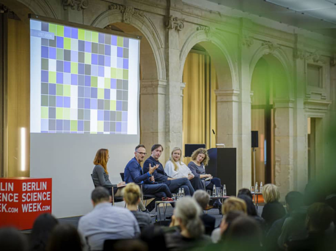 A panel of five speakers is seated on a stage, engaged in a discussion during an event. Behind them, a large screen displays a colorful grid of squares. The setting is formal, with an audience seated and attentively listening. The event is a part of "Berlin Science Week," as indicated by the signs on the stage. The venue is an elegant hall with stone arches, adding a sense of grandeur to the event.