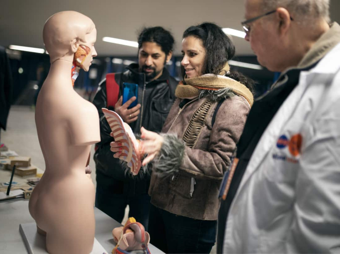 A group of individuals is gathered around a table displaying anatomical models. A woman in the center is holding and examining a detailed model of the human rib cage, while another person looks on and takes a photo with their phone. A man in a white coat, possibly a presenter or expert, stands nearby. The setting is indoors with modern lighting, creating a focused and informative atmosphere.