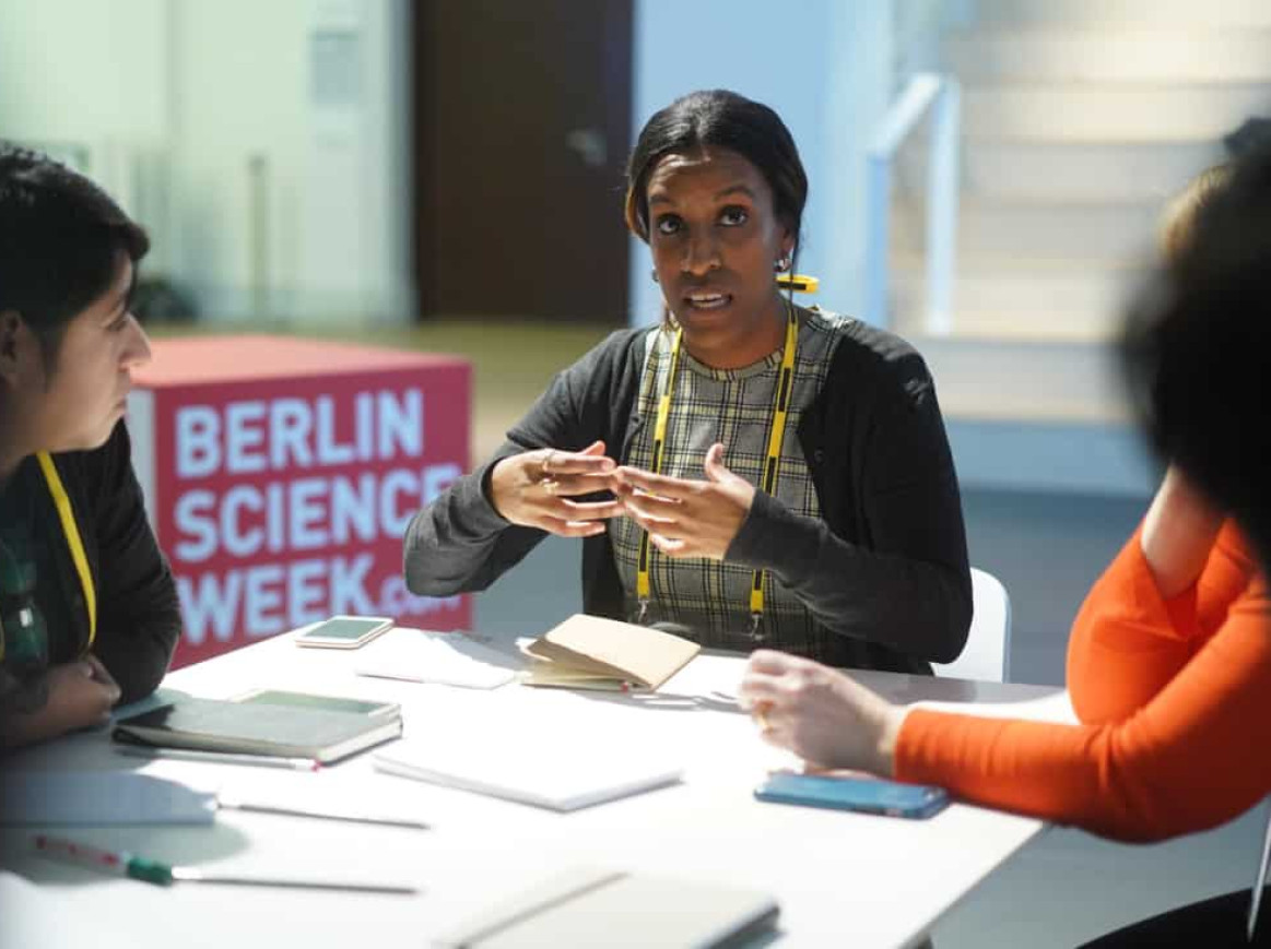 A woman speaks to two other people at a table during Berlin Science Week. The table is covered with notebooks and phones.