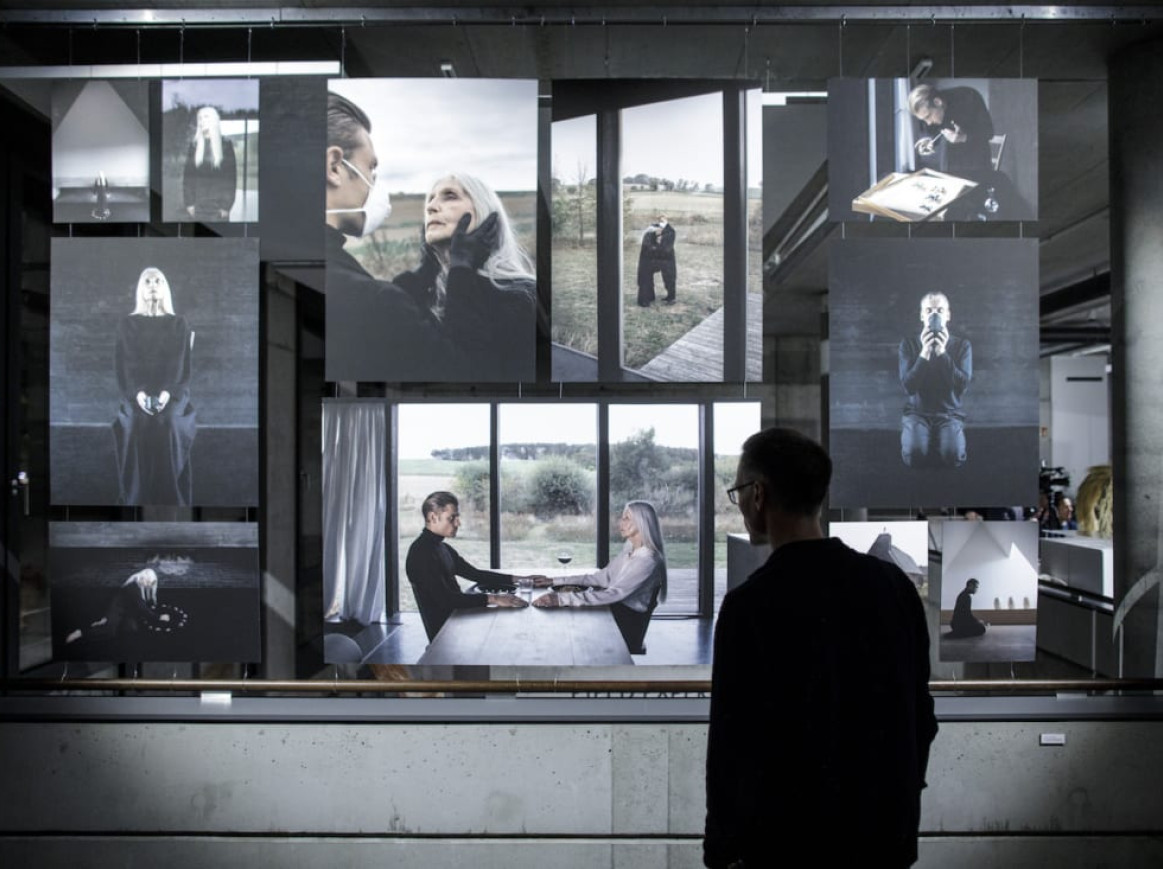 A person stands in front of a photo exhibition displaying several photographs. The photos show various scenes, including portraits of people, a person sitting at a table, and an outdoor landscape. The exhibition is in a modern, concrete building.