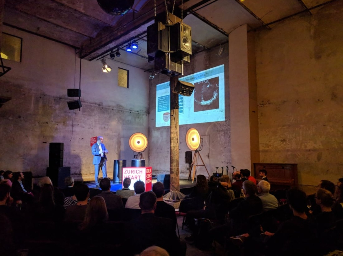 A speaker, dressed in a blue shirt, stands on a small stage in front of an audience, presenting a slide show in a dimly lit room with industrial decor. The screen displays medical images. A sign on stage reads "Zurich Heart." Two large circular lights are behind the speaker.