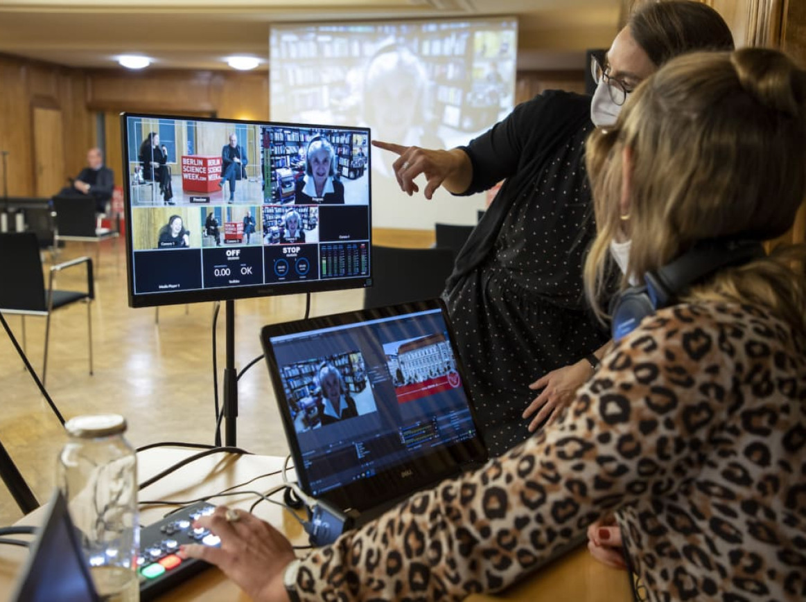 Two women work at a tech station with multiple screens and a laptop in a conference room. One woman is standing and pointing at the screen, explaining something, while the other is seated. Masks, microphones, and socially distanced seating are visible, indicating a hybrid event.