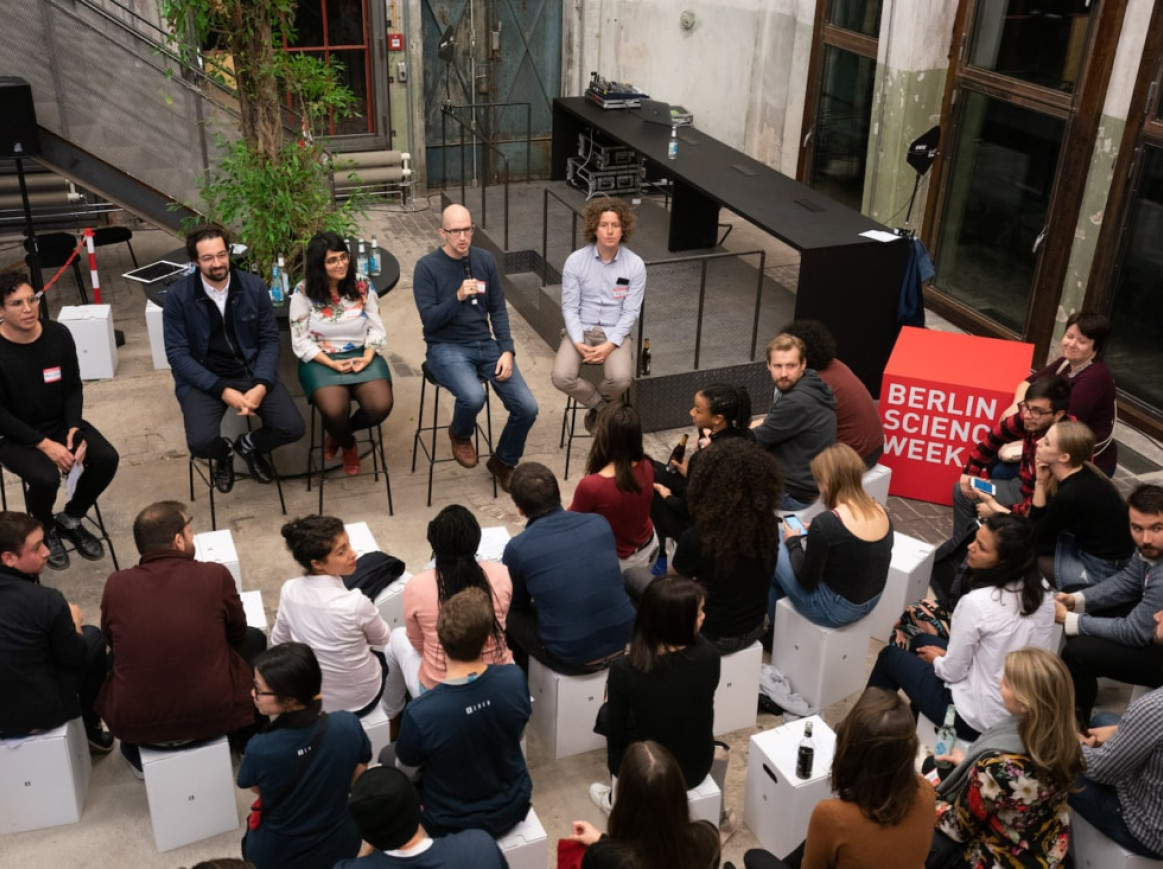 A group of people is attending a panel discussion during Berlin Science Week. The audience is seated facing five panelists who are seated in chairs. One of the panelists is speaking into a microphone. A sign that reads "Berlin Science Week" is visible on the right side.