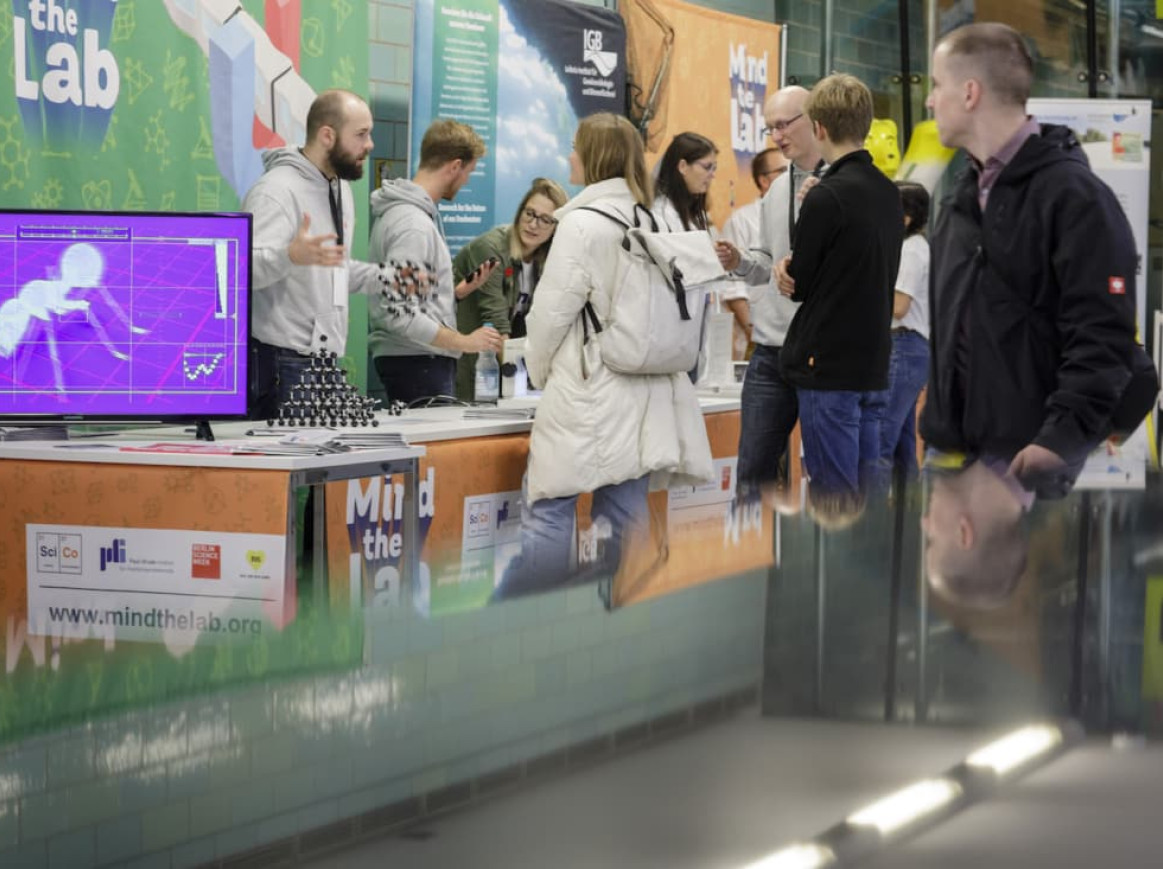 People engage in discussions at a science fair booth, titled "Mind the Lab." A screen displays a graphic of a human figure. The booth features posters and materials related to scientific research. Passersby observe and interact with the representatives at the booth.