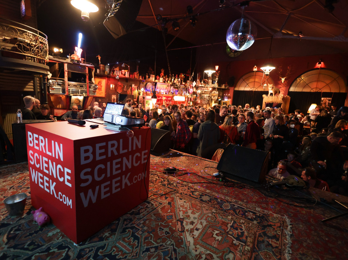 A shot taken from the stage into a bar full of people. On the stage there is a big red cube with the text "Berlin Science Week"