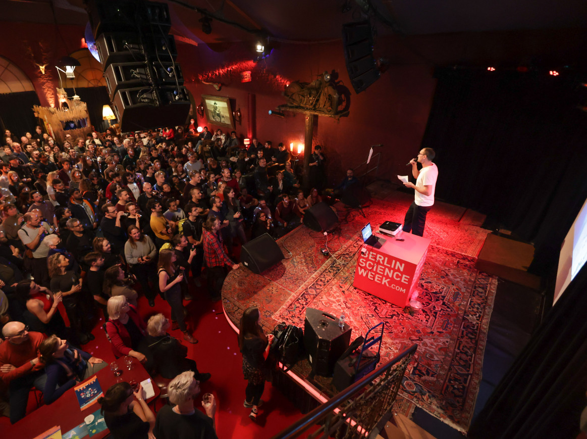 A shot from above of a bar full of people watching a person speaking on a stage that has a carpet and a red cube with the text "Berlin Science Week"