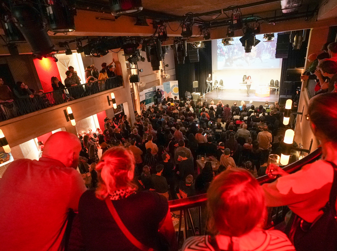 A red-lit room full with a balcony, full with a crowd looking at a stage