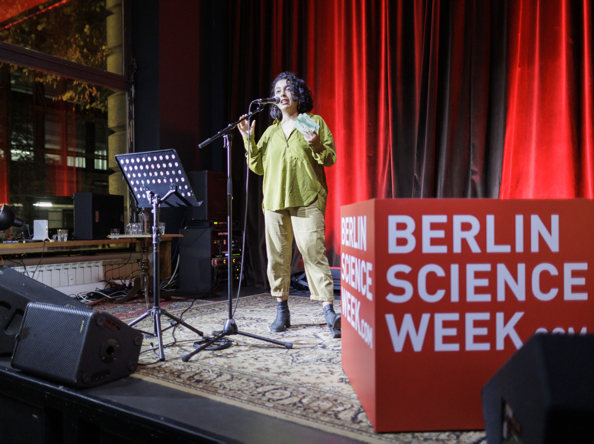 Person on stage speaking into a microphone next to a "Berlin Science Cube" cube