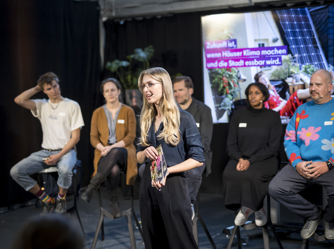 Shot of a woman speaking on stage, with other speakers behind her