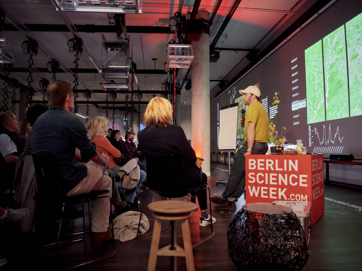 A man speaking to an audience while leaning on a "Berlin Science Week" cube