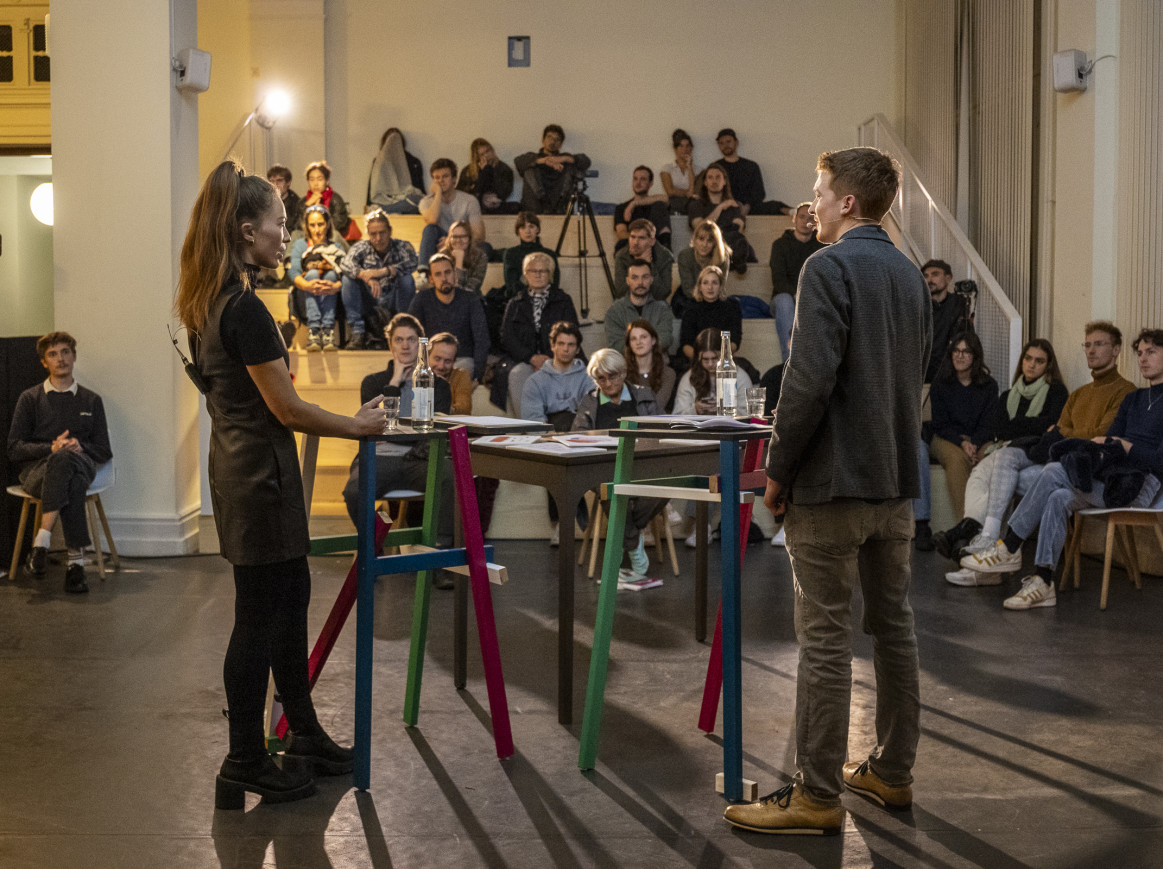 Two people standing talking in front of an audience indoors