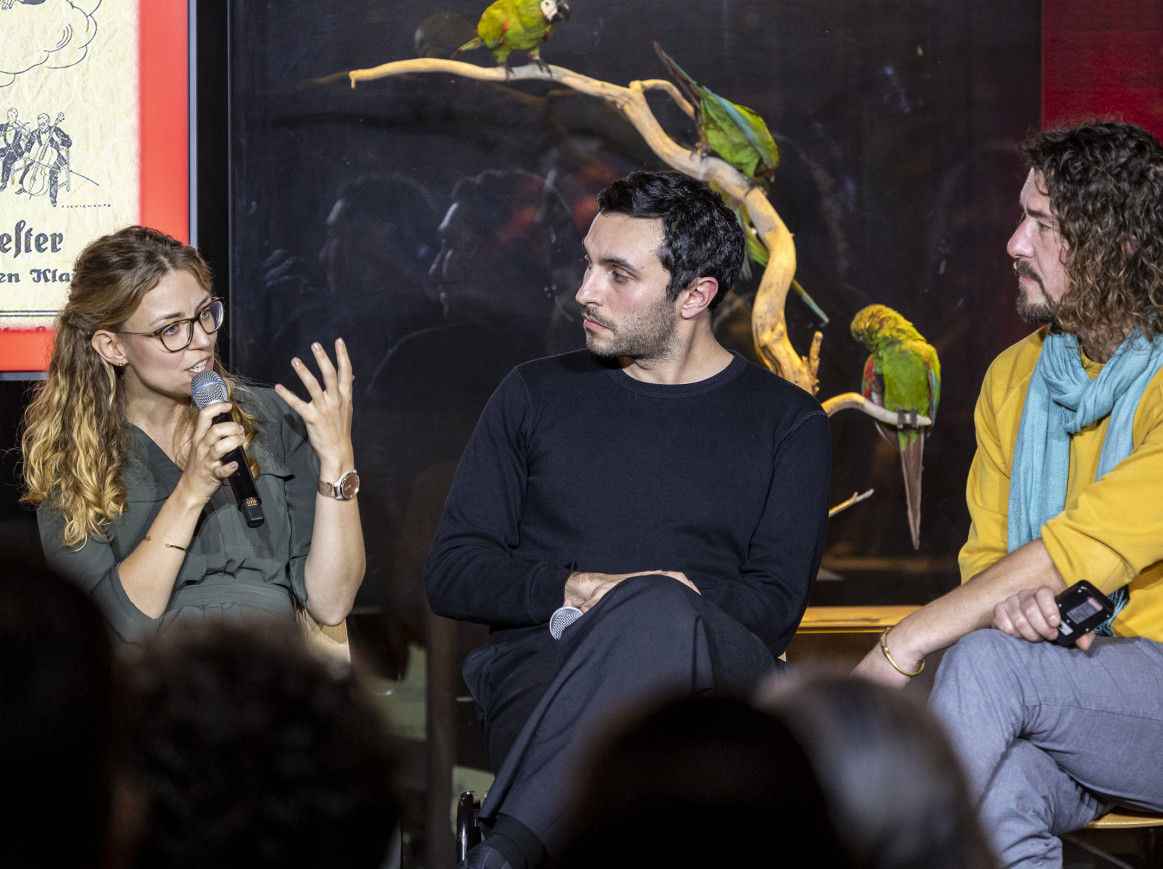 Two men and a woman talking on stage in front of a glass case with parrots