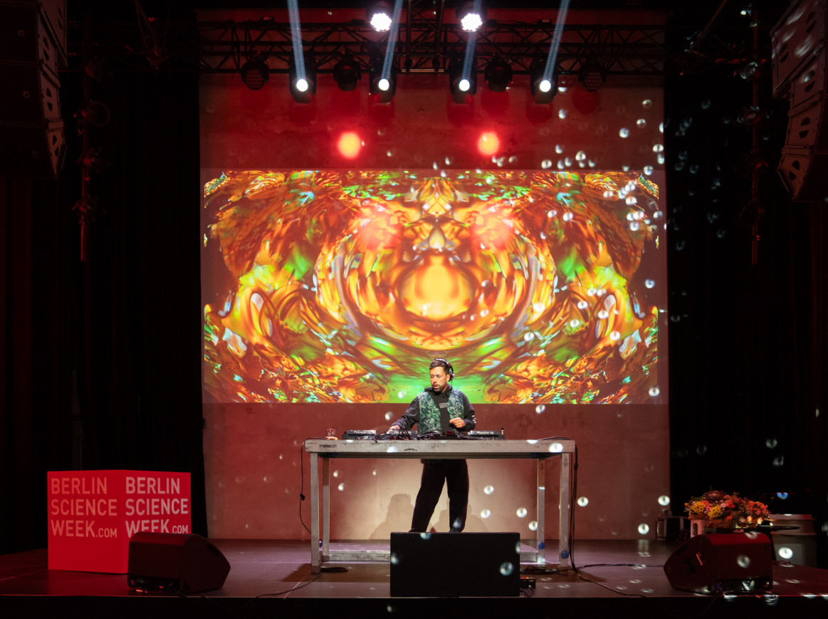 Science of Rave performing at the Berlin Science Week Closing Party with a DJ set and vibrant yellow visuals on stage.