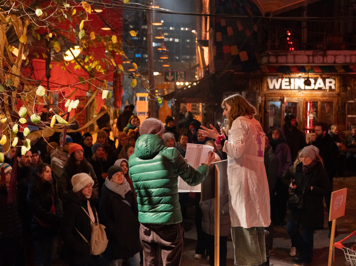 A Soapbox Science speaker standing on a small outdoor stage at the Holzmarkt 25 marketplace, surrounded by an engaged audience