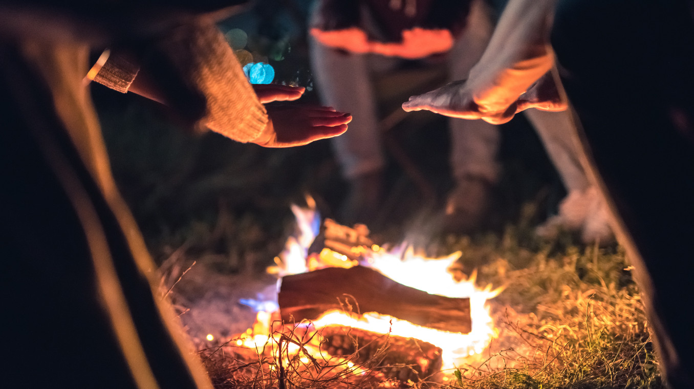 This image captures a cozy outdoor scene where people are gathered around a small campfire at night. Several hands are extended toward the fire, likely to warm up, as the flames crackle over the logs. The soft glow of the fire illuminates the surrounding area, casting warm light on the hands and nearby grass.