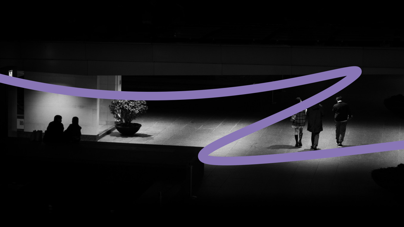 This image is showing a dimly lit urban scene at night. In the foreground, a wide walkway is illuminated by overhead lights. On the right side, three individuals walk away from the camera: two men and a woman, with the woman dressed in a checkered jacket or skirt, and the two men on either side of her. They walk towards a more shadowed area. To the left, under a bright light, two people sit on the floor, possibly talking, with a large potted plant next to them. The image has a minimalist, dark atmosphe