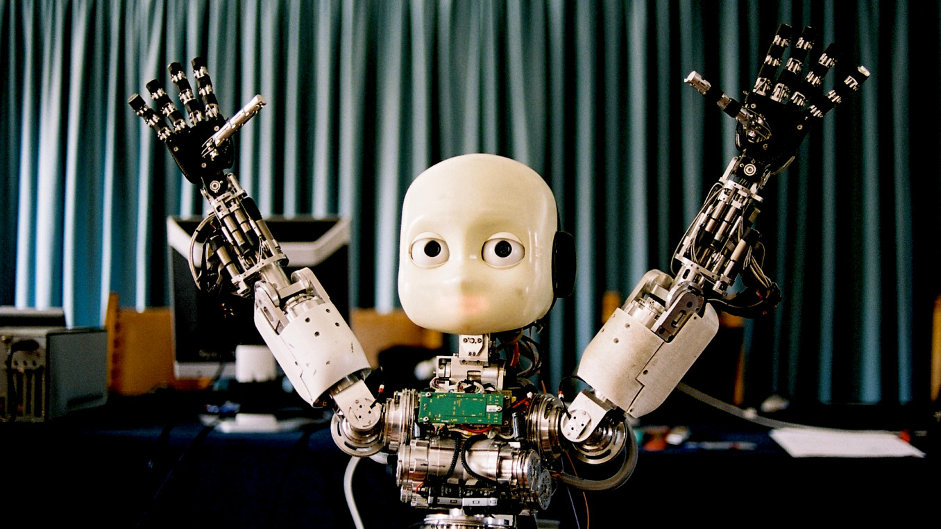 Das Bild zeigt einen humanoiden Roboter mit einem minimalistischen Gesicht, großen Augen und erhobenen Armen. Der Roboter hat komplexe mechanische Hände mit vielen beweglichen Teilen und ist teilweise freigelegt, sodass seine inneren elektronischen Komponenten sichtbar sind. Im Hintergrund hängt ein Vorhang, und es befinden sich Computer und technische Ausrüstung auf einem Tisch.